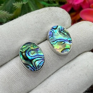 May include: Two oval-shaped abalone shell earrings with iridescent blue, green, and gold patterns. Each earring is set in a silver-colored metal frame. The earrings are displayed on a white surface.