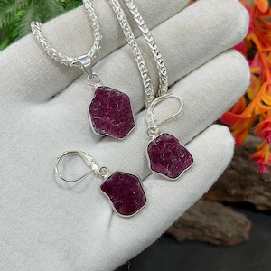 Op de afbeelding: Een sieradenset met een zilveren ketting met hanger en een paar oorbellen. Elk stuk toont een ruwe, onregelmatig gevormde, dieprode edelsteen. De edelstenen zijn gezet in zilveren zettingen, wat een opvallend contrast creëert.
