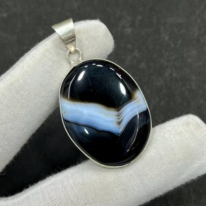May include: Oval pendant with a polished black and white agate stone, featuring a band of light blue. The stone is set in a silver-colored metal frame with a loop for a chain. The pendant is displayed on a white surface.