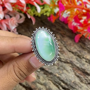 May include: Oval-shaped ring featuring a light green gemstone with a silver-colored, beaded border. The ring is held up by a hand, with a wooden surface in the background. The gemstone has a marbled appearance.