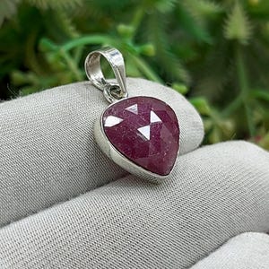May include: A silver pendant with a faceted, heart-shaped ruby gemstone. The ruby is a deep red color and is set in a silver bezel. The pendant has a simple, elegant design and is suitable for everyday wear.