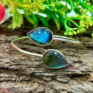 Può includere: Un bracciale a polsino argentato con due pietre di labradorite a forma di goccia. Le pietre mostrano sfumature iridescenti blu e verdi. Il bracciale è appoggiato su una superficie di legno marrone strutturata, con fogliame verde sullo sfondo.