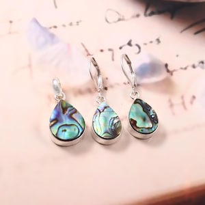 Könnte beinhalten: Schmuckset aus Abalone-Muscheln mit Anhänger und zwei Ohrringen. Die tropfenförmigen Abalone-Muscheln zeigen schillernde Blau-, Grün- und Lilatöne, gefasst in silberfarbenem Metall. Die Ohrringe haben Klappbügelverschlüsse. Der Schmuck wird auf einer hellrosa Oberfläche präsentiert.