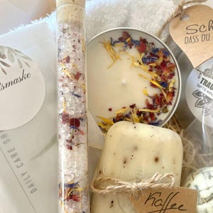 May include: A gift basket filled with bath and body products. The basket includes a white towel, a candle with dried flowers, a glass tube filled with bath salts, a bar of soap with a coffee scent, and a tag that says "Kaffee".