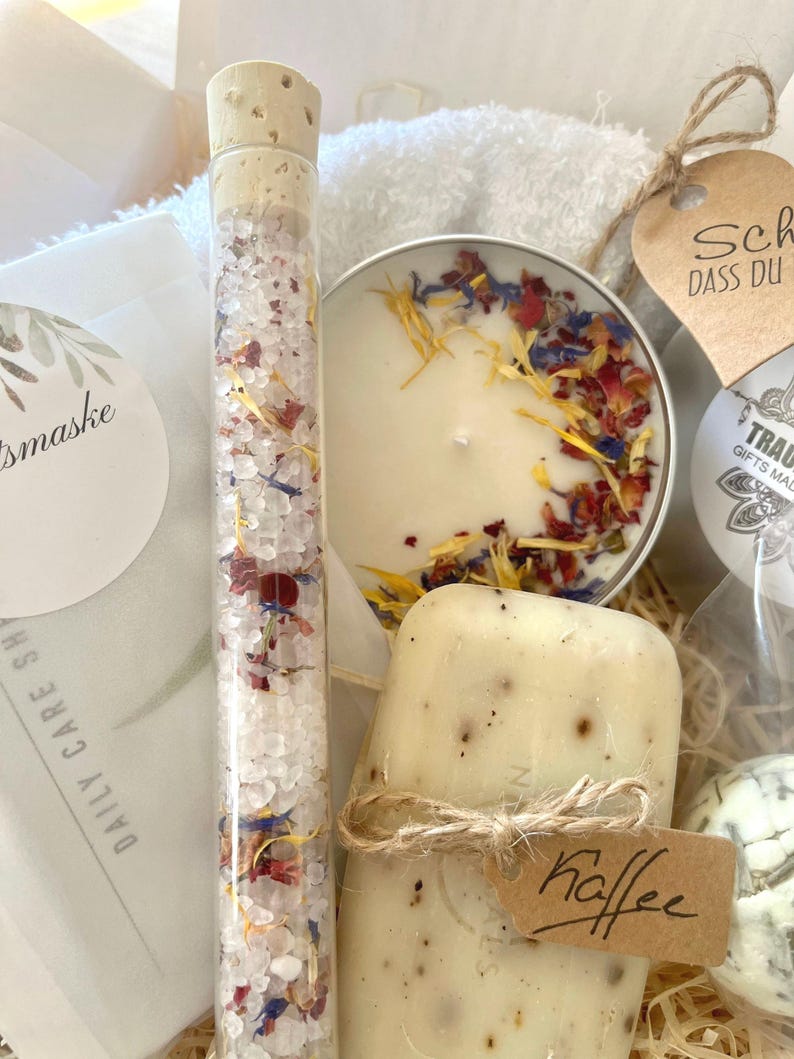 May include: A gift basket filled with bath and body products. The basket includes a white towel, a candle with dried flowers, a glass tube filled with bath salts, a bar of soap with a coffee scent, and a tag that says "Kaffee".