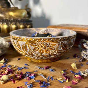 Könnte beinhalten: Ein geschnitzter Holz-Räucherofen mit einem floralen Design in Braun- und Goldtönen. Der Räucherofen ist mit Räucherwerk gefüllt und steht auf einer Holzoberfläche mit verstreuten getrockneten Blütenblättern.