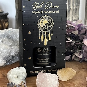Könnte beinhalten: Eine schwarze Schachtel mit "BOHO DREAMS" Myrrhe & Sandelholz Manifestations-Aromaöl. Die Schachtel hat goldene Akzente, darunter ein Traumfänger-Design und Sterne. Kristalle und getrockneter Lavendel sind ebenfalls zu sehen.