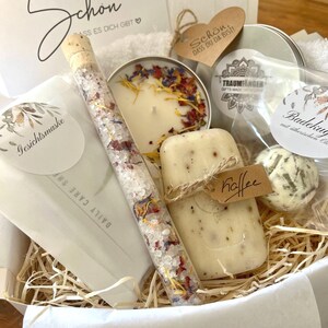 May include: A gift basket filled with bath and body products. The basket includes a candle, soap, bath salts, a bath bomb, and a small bottle of bath oil. The basket is lined with shredded paper and has a white paper liner. The basket is decorated with a small tag that says "Schon, dass du da bist!" which translates to "It's nice that you're here!"