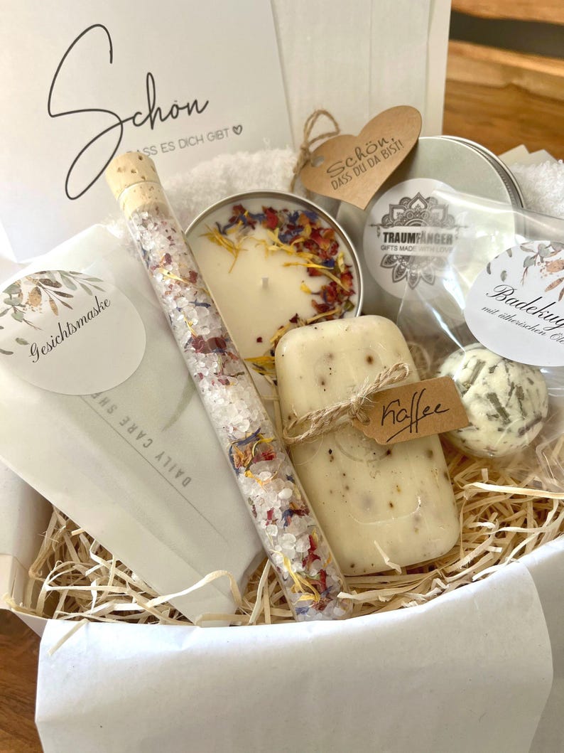 May include: A gift basket filled with bath and body products. The basket includes a candle, soap, bath salts, a bath bomb, and a small bottle of bath oil. The basket is lined with shredded paper and has a white paper liner. The basket is decorated with a small tag that says "Schon, dass du da bist!" which translates to "It's nice that you're here!"