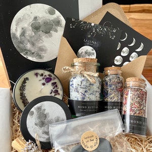 Könnte beinhalten: Ein Ritual-Set mit einer Kerze mit violetten Steinen und Lavendel, drei Glasgefäßen mit Korkstopfen gefüllt mit getrockneten Kräutern und Salzen, einem Klarsichtbeutel mit zwei dunkelgrauen Scheiben sowie Karten mit Mondmotiven und dem Text MOND RITUAL.
