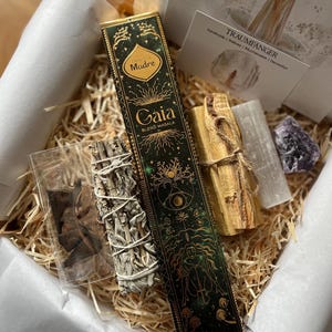 Könnte beinhalten: Eine Geschenkbox gefüllt mit natürlichen Wellness-Artikeln. Die Box enthält eine grün-goldene Räucherstäbchenbox mit dem Wort "Gaia", ein Bündel weißen Salbeis, ein Stück Selenit, ein Stück Palo Santo und einen kleinen Amethystkristall. Die Box ist mit weißem Papier und Stroh ausgekleidet.