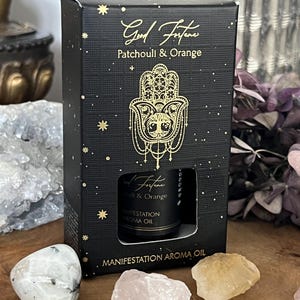 Könnte beinhalten: Schwarze Schachtel mit goldenem Text und einem Hamsa-Hand-Design. Auf der Schachtel steht "Good Fortune Patchouli & Orange" und sie enthält eine Flasche "Manifestation Aroma Oil". Kristalle und Trockenblumen sind ebenfalls zu sehen.