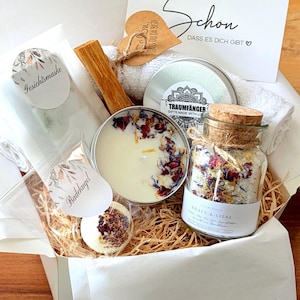 Könnte beinhalten: Eine Geschenkbox mit einem weißen Handtuch, einer Kerze in einer Metalldose mit getrockneten Blütenblättern, einem Glas mit Badesalz und Korkverschluss, einer Badekugel, einem Holzstab und einer Karte mit dem Text Schon DASS ES DICH GIBT.