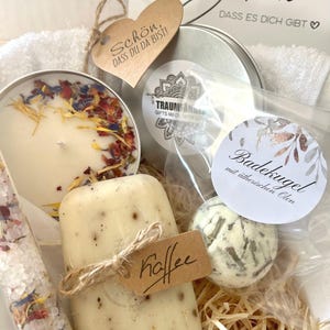 May include: A gift basket filled with bath and body products. The basket includes a candle, bath bomb, soap, bath salts, and a small towel. The basket is decorated with a brown heart-shaped tag that says "Sch&ouml;n, dass du da bist!" and a small brown tag that says "Kaffee".