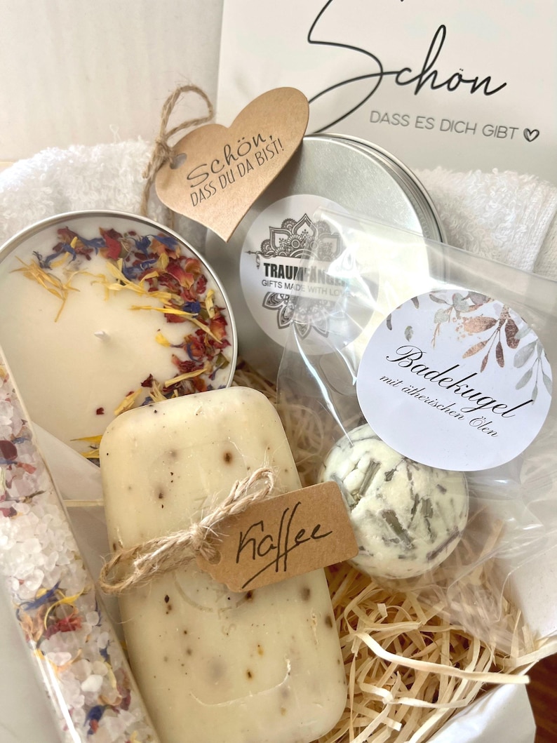 May include: A gift basket filled with bath and body products. The basket includes a candle, bath bomb, soap, bath salts, and a small towel. The basket is decorated with a brown heart-shaped tag that says "Sch&ouml;n, dass du da bist!" and a small brown tag that says "Kaffee".