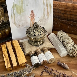 Könnte beinhalten: Ein Räucherset mit verschiedenen Artikeln, darunter ein Salbei-Räucherstäbchen, ein Bergkristall, ein kleiner Räucherofen, drei kleine Glasflaschen mit Etiketten und ein Bund getrockneter Lavendel. Das Set ist auf einer Holzoberfläche angeordnet.