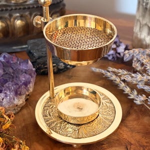 Könnte beinhalten: Ein goldfarbener Metall-Ölbrenner mit einer runden Basis mit geprägtem Blumenmuster, einem zentralen Kerzenhalter und einer verstellbaren oberen Schale mit perforiertem Sieb zum Erhitzen von Ölen oder Harzen, gehalten von einem vertikalen Metallstab.