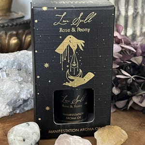 Könnte beinhalten: Eine schwarze Schachtel mit goldenen Buchstaben und Illustrationen, die eine kleine Glasflasche enthält. Auf der Schachtel steht "Love Spell Rose & Peony" und "Manifestation Aroma Oil". Mehrere Kristalle, darunter Rosenquarz, sind vor der Schachtel angeordnet.