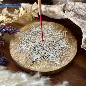 Könnte beinhalten: Ein runder, hölzerner Räucherstäbchenhalter mit einem weißen Mandala-Design und goldenen Akzenten. Der Halter hat in der Mitte ein kleines Loch für ein Räucherstäbchen.