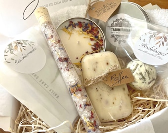 May include: A gift basket filled with bath and body products, including a candle, bath salts, soap, and a bath bomb. The basket is lined with shredded paper and has a white tissue paper lining. The basket is decorated with a tag that says "Schon, dass du da bist!" which translates to "It's nice that you're here!" in English.
