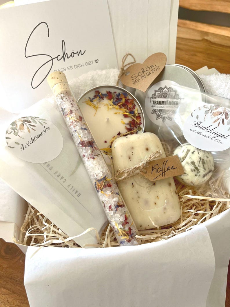 May include: A gift basket filled with bath and body products, including a candle, bath salts, soap, and a bath bomb. The basket is lined with shredded paper and has a white tissue paper lining. The basket is decorated with a tag that says "Schon, dass du da bist!" which translates to "It's nice that you're here!" in English.