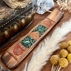 Könnte beinhalten: Holz-Räucherstäbchenhalter mit einem keltischen Knotendesign und grünen Aventurin-Kristallen. Der Halter steht auf einer Holzoberfläche mit getrockneten Blumen und einer weißen Feder.