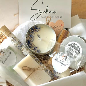 Könnte beinhalten: Eine Geschenkbox mit einer weißen Kerze in einer Dose mit blauen Blütenblättern, einem mit Kordel gebundenen Seifenstück, einem Glasröhrchen mit Badesalz, einer Badekugel in einer Klarsichttüte, einem Holzherz und einer Karte mit dem Text Schon dass es dich gibt.