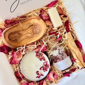 Könnte beinhalten: Eine Geschenkbox gefüllt mit Holzwolle und getrockneten Rosenblättern, die eine ovale Schale aus Olivenholz, ein rechteckiges weißes Seifenstück, eine runde weiße Kerze mit Rosenblättern und ein Glas mit Badesalz mit der Aufschrift KRAFT & LIEBE enthält.