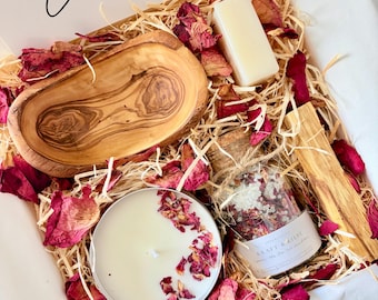 May include: A gift box filled with wood shavings and dried rose petals containing an oval olive wood dish, a rectangular white soap bar, a round white candle topped with rose petals, and a glass jar of bath salts labelled KRAFT & LIEBE.