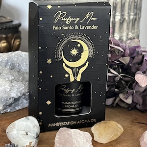 Könnte beinhalten: Eine schwarze Schachtel mit goldenen Sternen und dem Text "Purifying Moon Palo Santo & Lavender" enthält eine kleine Flasche "Manifestation Aroma Oil". Kristalle und getrockneter Lavendel liegen auf einer Holzoberfläche.