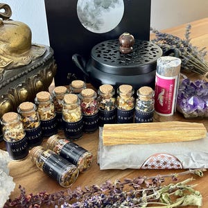 Könnte beinhalten: Eine Sammlung spiritueller Artikel, darunter kleine Glasgläser mit Korken, ein schwarzer Räuchergefäß, ein Buch mit Mondgrafik, getrocknete Kräuter, Kristalle und ein Palo Santo-Stab. Die Gläser haben deutsche Etiketten.