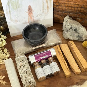 Könnte beinhalten: Ein Ritualset bestehend aus einer schwarzen Steinschale, einer Packung Räucherkohle mit der Aufschrift Kohletabletten, drei kleinen Glasfläschchen mit den Etiketten Merit, Styrax und Myrrhe, einem weißen Salbei-Bündel, einem Selenitstab und drei Palo-Santo-Hölzern.