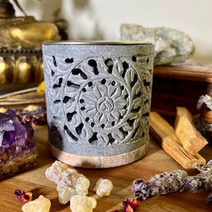 Könnte beinhalten: Grauer Stein-Räucherofen mit aufwendig geschnitzten floralen Mustern. Der Ofen hat eine runde Oberseite und einen flachen Boden. Er steht auf einer Holzoberfläche mit anderen Räucherutensilien.