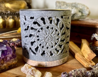 May include: Gray stone incense burner with intricate carved floral designs. The burner has a round top and a flat base. It is sitting on a wooden surface with other incense related items.