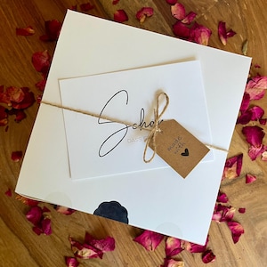 May include: A white box with a brown tag that says "made with" and a heart. The box is tied with twine and has a white card on top with the text "Schon" and "DASS F". The box is surrounded by pink rose petals on a wooden surface.
