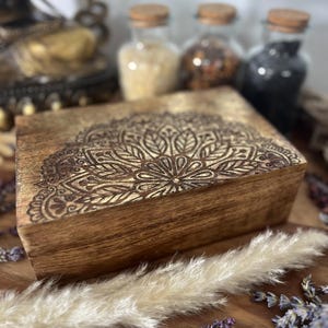 Könnte beinhalten: Eine braune Holzbox mit einem eingravierten Mandala-Muster auf dem Deckel. Die Box hat ein goldfarbenes Design. Der Hintergrund umfasst Gläser mit Korkdeckeln, Trockenblumen und dekorative Elemente, die eine Boho-Ästhetik schaffen.