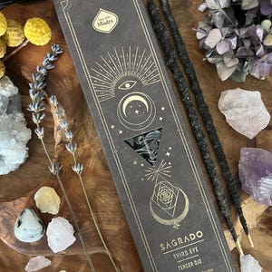 Könnte beinhalten: Eine dunkelbraune Schachtel mit Räucherstäbchen mit goldenen Himmelsmustern und den Worten "SAGRADO THIRD EYE" darauf. Zwei schwarze Räucherstäbchen liegen neben der Schachtel. Das Bild enthält auch getrockneten Lavendel, Kristalle und getrocknete Blumen.