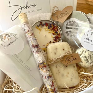 May include: A gift basket filled with bath and body products. The basket includes a candle, bath salts, soap, and a bath bomb. The basket is decorated with a tag that says "Sch&ouml;n. Dass du da bist!" which translates to "Beautiful. That you are here!"