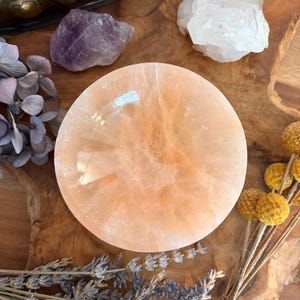 Könnte beinhalten: Eine pfirsichfarbene, runde Kristallschale liegt auf einer Holzoberfläche, umgeben von Amethyst- und Quarz-Kristallen, getrocknetem Lavendel und gelben Blumen. Die Schale hat eine glatte, polierte Oberfläche. Die Anordnung deutet auf ein Thema natürlicher Elemente und spiritueller Praktiken hin.