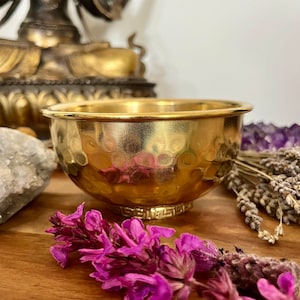 Könnte beinhalten: Eine goldene Metallschale mit gehämmerter Oberfläche. Die Schale hat einen dekorativen Rand um den Rand. Die Schale steht auf einer Holzoberfläche mit violetten Blumen.