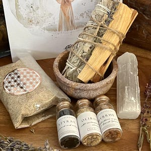 Könnte beinhalten: Ein Set aus drei kleinen Glasflaschen mit den Etiketten "Styrax", "Olibanum" und "Myrrhe", die Harze zum Verbrennen enthalten. Die Flaschen stehen auf einer Holzoberfläche mit einem Salbei-Räucherstäbchen, einem Palo-Santo-Stäbchen und einem klaren Quarzkristall.