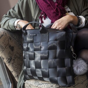 Könnte beinhalten: Eine schwarze Tragetasche mit einem gewebten Design aus Sicherheitsgurtmaterial. Die Tasche hat einen oberen Griff und einen Reißverschluss. Ein flauschiger grauer Pompon hängt am Reißverschluss. Die Tasche wird von einer Person gehalten, die auf einem Stuhl sitzt.