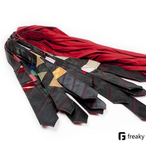 Può includere: Una collezione di cravatte nere con vari disegni patchwork colorati e cuciture rosse. Le cravatte sono disposte con un vivace tessuto rosso sullo sfondo, il tutto su uno sfondo bianco. Le cravatte sono di Freaky Goods.