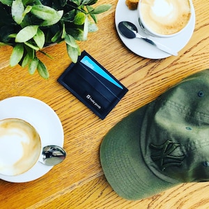 Könnte beinhalten: Draufsicht auf einen Holztisch mit einem schwarzen Kartenhalter, einer grünen Baseballkappe und zwei Tassen Kaffee. Der Kartenhalter hat eine blaue Karte und den Text "freaky goods". Die Baseballkappe hat ein dunkelgrünes Emblem.