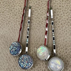 May include: Three silver hair clips with decorative round tops. Two clips have iridescent glitter tops and one clip has a white iridescent top.