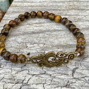 May include: A beaded bracelet featuring round, brown tiger eye stones. The bracelet has a decorative, antique brass-colored centerpiece with an ornate, swirling design. The bracelet is strung on an elastic band.