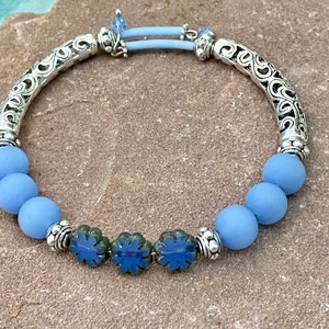 Puede incluir: Una pulsera azul claro con cuentas redondas mate y tres cuentas azules en forma de flor. La pulsera tiene detalles plateados en forma de tubo ornamentado y una banda elástica azul claro. Una joya delicada.