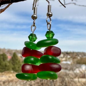 May include: A pair of handmade earrings featuring alternating red and green sea glass pieces. The earrings are suspended from silver-colored fish hook ear wires and include a green faceted bead at the top. The earrings are displayed against a blurred outdoor background.