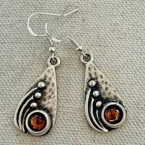 May include: A pair of silver-toned drop earrings featuring a teardrop-shaped pendant with a hammered texture. Each earring displays three small raised spheres and a single round, translucent amber-colored stone set within a circular frame.