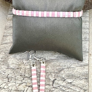 Könnte beinhalten: Ein Armband und Ohrringe mit einem rosa-weiß gestreiften Muster. Der Schmuck wird auf einem grauen Kissen präsentiert. Die Ohrringe haben ein vertikales Streifenmuster, und das Armband ist horizontal. Der Schmuck besteht aus kleinen rechteckigen Perlen.